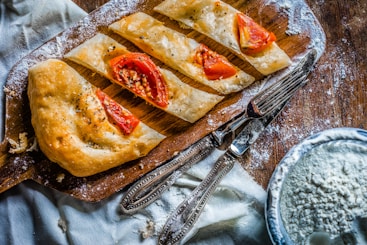 A wooden cutting board displays slices of baked bread adorned with tomato segments and herbs. The surface is dusted with flour, suggesting a baking or cooking activity. Beside the board, there are utensils and a bowl containing a powdery substance, likely more flour, on a rustic wooden table.