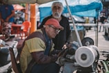 Two men are engaged in a mechanical task outdoors, with one wearing glasses and a red cap while examining a machine. The setting appears to be a busy outdoor market or workshop area, with bright colors and a mix of objects in the background.