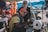 Two men are engaged in a mechanical task outdoors, with one wearing glasses and a red cap while examining a machine. The setting appears to be a busy outdoor market or workshop area, with bright colors and a mix of objects in the background.