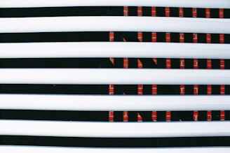 Horizontal white blinds partially covering a digital display or screen with a repeating pattern of red lights against a dark background.
