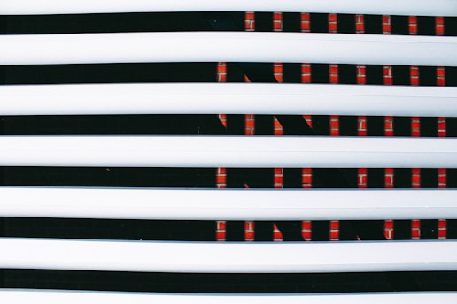 Horizontal white blinds partially covering a digital display or screen with a repeating pattern of red lights against a dark background.