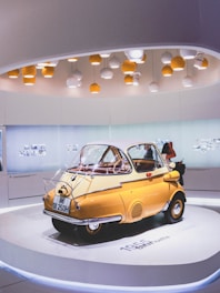 A classic yellow microcar is displayed on a circular platform in a modern exhibition room. The car, with a compact and unique design, features a front door opening and sits beneath a ceiling decorated with numerous spherical pendant lights in white and orange. The backdrop includes minimalist white walls with subtle artwork or photographs, contributing to a clean and modern ambiance.