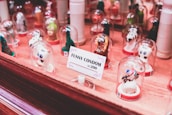 A collection of novelty condoms shaped like various animals and characters are displayed under small glass domes. They are arranged on a wooden surface with a sign that reads 'FUNNY CONDOM kc 200' in the foreground. The condoms have playful designs with vivid colors.