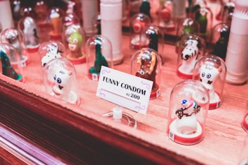 A collection of novelty condoms shaped like various animals and characters are displayed under small glass domes. They are arranged on a wooden surface with a sign that reads 'FUNNY CONDOM kc 200' in the foreground. The condoms have playful designs with vivid colors.