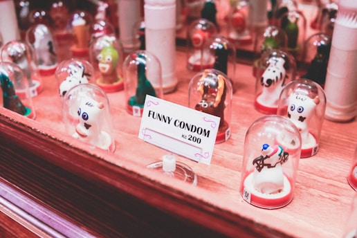 A collection of novelty condoms shaped like various animals and characters are displayed under small glass domes. They are arranged on a wooden surface with a sign that reads 'FUNNY CONDOM kc 200' in the foreground. The condoms have playful designs with vivid colors.