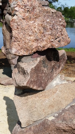 Several large, rough stones are stacked upon each other in an outdoor setting. The stones have various shades of brown and gray, with a textured, jagged surface. In the background, there is lush greenery and a glimpse of a body of water.