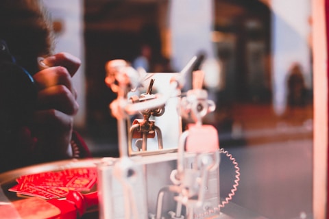 A close-up view of a mechanical object with metallic parts and gears, placed near a reflective surface. There is a hand in the background, partially visible, suggesting interaction or adjustment of the machinery. The scene is set indoors, featuring warm lighting and a blurred background.
