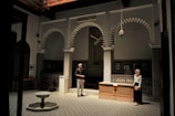 A beautifully designed interior space featuring intricate tile work and classic Islamic architecture. The room has patterned walls and floors, with three grand arches and traditional lanterns hanging from the ceiling. Two people are present, a man and a woman, engaged in conversation. On the floor, there is a small fountain, and the ambient lighting creates a serene and contemplative atmosphere.