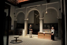 A beautifully designed interior space featuring intricate tile work and classic Islamic architecture. The room has patterned walls and floors, with three grand arches and traditional lanterns hanging from the ceiling. Two people are present, a man and a woman, engaged in conversation. On the floor, there is a small fountain, and the ambient lighting creates a serene and contemplative atmosphere.
