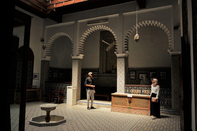 A beautifully designed interior space featuring intricate tile work and classic Islamic architecture. The room has patterned walls and floors, with three grand arches and traditional lanterns hanging from the ceiling. Two people are present, a man and a woman, engaged in conversation. On the floor, there is a small fountain, and the ambient lighting creates a serene and contemplative atmosphere.