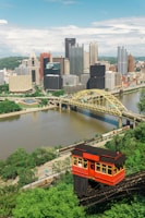 A vibrant cityscape with a classic red cable car ascending a steep greenery-laden track in the foreground. The city skyline features a variety of tall, modern skyscrapers and historic-looking buildings. A river flows through the city with a distinctive yellow bridge crossing over it.