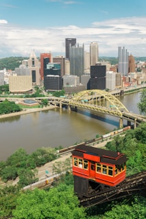 A vibrant cityscape with a classic red cable car ascending a steep greenery-laden track in the foreground. The city skyline features a variety of tall, modern skyscrapers and historic-looking buildings. A river flows through the city with a distinctive yellow bridge crossing over it.