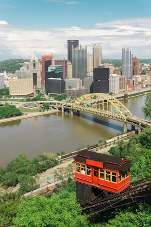 A vibrant cityscape with a classic red cable car ascending a steep greenery-laden track in the foreground. The city skyline features a variety of tall, modern skyscrapers and historic-looking buildings. A river flows through the city with a distinctive yellow bridge crossing over it.