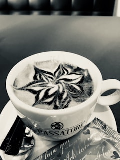 A cup of coffee with intricate latte art on the surface, displaying a star-like pattern. The cup is placed on a saucer with a spoon and a wrapped biscuit beside it. The background appears to be a seating area, possibly in a café.