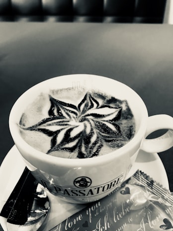 A cup of coffee with intricate latte art on the surface, displaying a star-like pattern. The cup is placed on a saucer with a spoon and a wrapped biscuit beside it. The background appears to be a seating area, possibly in a café.