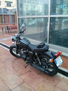 A black motorcycle is parked on a wet, tiled pavement near a glass building. The motorcycle has a license plate with the number 9323 JLK. The surrounding environment features some buildings, benches, and a parked car visible through the glass reflection.