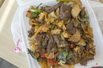 A takeout container filled with a savory dish that includes wide rice noodles, pieces of beef, scrambled egg fragments, green and red pepper slices, and fresh basil leaves.
