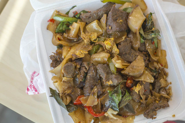 A takeout container filled with a savory dish that includes wide rice noodles, pieces of beef, scrambled egg fragments, green and red pepper slices, and fresh basil leaves.