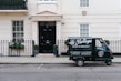 A small delivery truck with the branding 'La Bottega' is parked on a city street in front of a classic, beige townhouse. The vehicle is painted in dark green with white lettering and is positioned next to an iron fence with two potted plants on either side of a black door. There are two windows with white frames and small decorative planters underneath.
