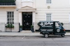A small delivery truck with the branding 'La Bottega' is parked on a city street in front of a classic, beige townhouse. The vehicle is painted in dark green with white lettering and is positioned next to an iron fence with two potted plants on either side of a black door. There are two windows with white frames and small decorative planters underneath.