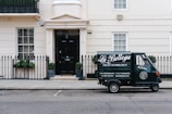 A small delivery truck with the branding 'La Bottega' is parked on a city street in front of a classic, beige townhouse. The vehicle is painted in dark green with white lettering and is positioned next to an iron fence with two potted plants on either side of a black door. There are two windows with white frames and small decorative planters underneath.