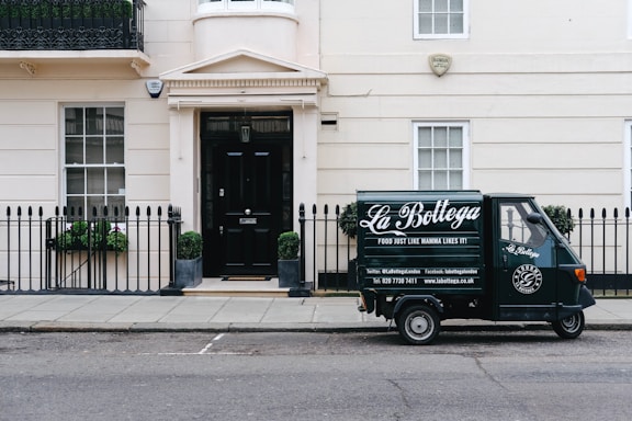 A small delivery truck with the branding 'La Bottega' is parked on a city street in front of a classic, beige townhouse. The vehicle is painted in dark green with white lettering and is positioned next to an iron fence with two potted plants on either side of a black door. There are two windows with white frames and small decorative planters underneath.