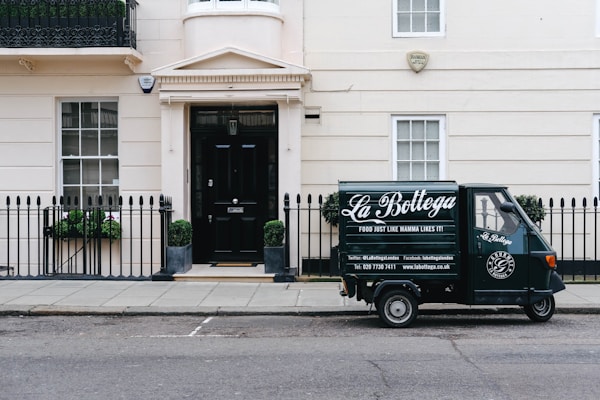 A small delivery truck with the branding 'La Bottega' is parked on a city street in front of a classic, beige townhouse. The vehicle is painted in dark green with white lettering and is positioned next to an iron fence with two potted plants on either side of a black door. There are two windows with white frames and small decorative planters underneath.