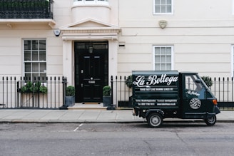 A small delivery truck with the branding 'La Bottega' is parked on a city street in front of a classic, beige townhouse. The vehicle is painted in dark green with white lettering and is positioned next to an iron fence with two potted plants on either side of a black door. There are two windows with white frames and small decorative planters underneath.