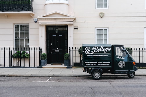 A small delivery truck with the branding 'La Bottega' is parked on a city street in front of a classic, beige townhouse. The vehicle is painted in dark green with white lettering and is positioned next to an iron fence with two potted plants on either side of a black door. There are two windows with white frames and small decorative planters underneath.