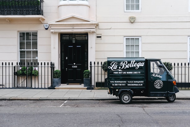 A small delivery truck with the branding 'La Bottega' is parked on a city street in front of a classic, beige townhouse. The vehicle is painted in dark green with white lettering and is positioned next to an iron fence with two potted plants on either side of a black door. There are two windows with white frames and small decorative planters underneath.