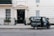 A small delivery truck with the branding 'La Bottega' is parked on a city street in front of a classic, beige townhouse. The vehicle is painted in dark green with white lettering and is positioned next to an iron fence with two potted plants on either side of a black door. There are two windows with white frames and small decorative planters underneath.