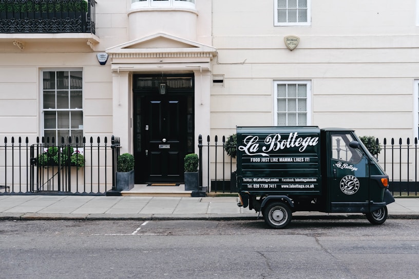 A small delivery truck with the branding 'La Bottega' is parked on a city street in front of a classic, beige townhouse. The vehicle is painted in dark green with white lettering and is positioned next to an iron fence with two potted plants on either side of a black door. There are two windows with white frames and small decorative planters underneath.