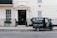 A small delivery truck with the branding 'La Bottega' is parked on a city street in front of a classic, beige townhouse. The vehicle is painted in dark green with white lettering and is positioned next to an iron fence with two potted plants on either side of a black door. There are two windows with white frames and small decorative planters underneath.