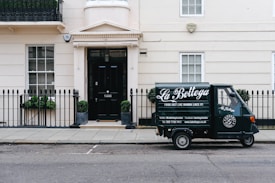 A small delivery truck with the branding 'La Bottega' is parked on a city street in front of a classic, beige townhouse. The vehicle is painted in dark green with white lettering and is positioned next to an iron fence with two potted plants on either side of a black door. There are two windows with white frames and small decorative planters underneath.