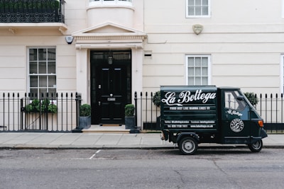 A small delivery truck with the branding 'La Bottega' is parked on a city street in front of a classic, beige townhouse. The vehicle is painted in dark green with white lettering and is positioned next to an iron fence with two potted plants on either side of a black door. There are two windows with white frames and small decorative planters underneath.
