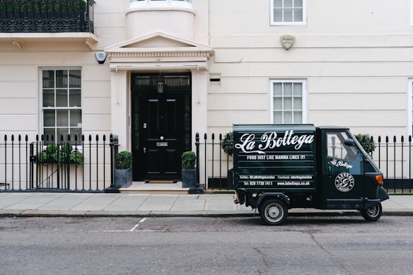 A small delivery truck with the branding 'La Bottega' is parked on a city street in front of a classic, beige townhouse. The vehicle is painted in dark green with white lettering and is positioned next to an iron fence with two potted plants on either side of a black door. There are two windows with white frames and small decorative planters underneath.