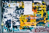 A weathered bulletin board covered with a chaotic mix of torn posters, flyers, and vibrant graffiti. Text in various languages is partially visible. Yellow, white, and blue are prominent among other overlapping layers of paper and paint.
