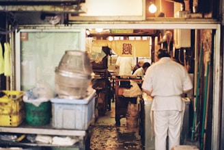 A cozy and cluttered interior of what appears to be a small restaurant kitchen. Several people dressed in white uniforms are busy preparing food. The space is filled with various kitchen equipment and storage containers. The warm lighting creates an intimate and inviting atmosphere, with a traditional-style banner hanging in the background.