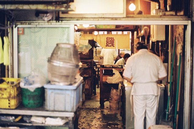 A cozy and cluttered interior of what appears to be a small restaurant kitchen. Several people dressed in white uniforms are busy preparing food. The space is filled with various kitchen equipment and storage containers. The warm lighting creates an intimate and inviting atmosphere, with a traditional-style banner hanging in the background.