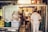 A cozy and cluttered interior of what appears to be a small restaurant kitchen. Several people dressed in white uniforms are busy preparing food. The space is filled with various kitchen equipment and storage containers. The warm lighting creates an intimate and inviting atmosphere, with a traditional-style banner hanging in the background.