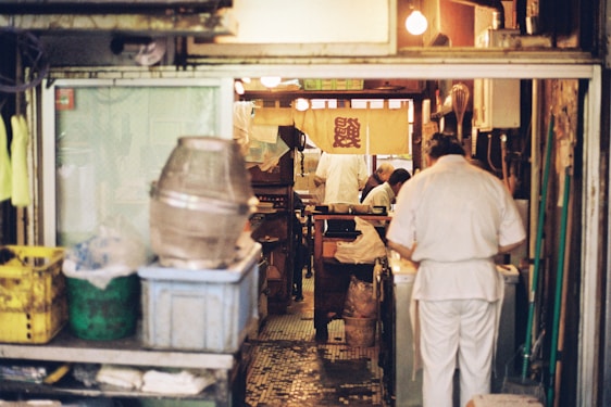 A cozy and cluttered interior of what appears to be a small restaurant kitchen. Several people dressed in white uniforms are busy preparing food. The space is filled with various kitchen equipment and storage containers. The warm lighting creates an intimate and inviting atmosphere, with a traditional-style banner hanging in the background.