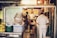 A cozy and cluttered interior of what appears to be a small restaurant kitchen. Several people dressed in white uniforms are busy preparing food. The space is filled with various kitchen equipment and storage containers. The warm lighting creates an intimate and inviting atmosphere, with a traditional-style banner hanging in the background.