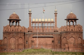 A historic red sandstone fort with impressive architectural features, including large domes and arched structures. The fort is adorned with an Indian flag flying on top, indicating its cultural significance.