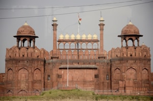 A historic red sandstone fort with impressive architectural features, including large domes and arched structures. The fort is adorned with an Indian flag flying on top, indicating its cultural significance.