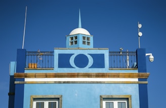 The image features the upper part of a building with a deep blue facade. A round, white dome with a small windowed structure sits on top, and a striking geometric design is displayed prominently on the wall below. Antennas and surveillance cameras are mounted on the rooftop, alongside decorative metal railing.