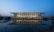 A modern architectural building is seen during twilight. It features a large, glass-fronted facade with steel framework, reflecting elegantly on a calm body of water in the foreground. The surroundings include landscaped areas and soft lighting, contributing to a serene ambiance.