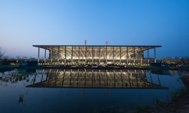 A modern architectural building is seen during twilight. It features a large, glass-fronted facade with steel framework, reflecting elegantly on a calm body of water in the foreground. The surroundings include landscaped areas and soft lighting, contributing to a serene ambiance.