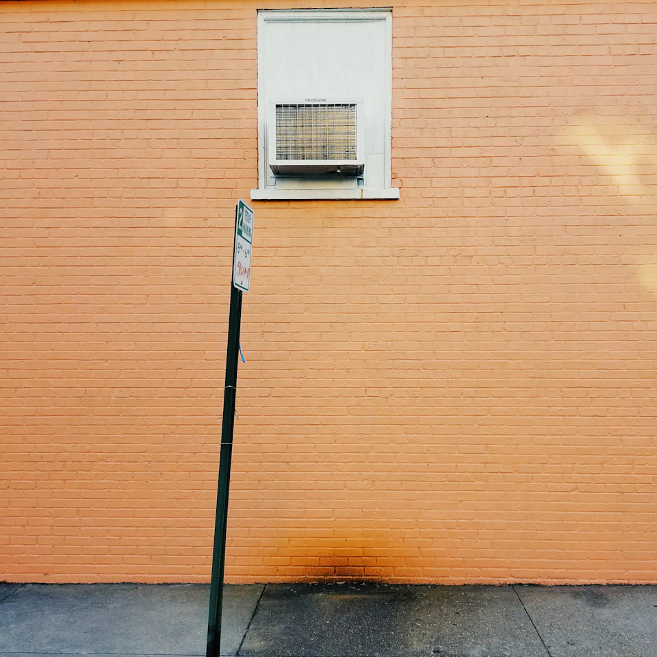 The image shows a brick wall with an orange hue, and a green street sign leaning against it on the left side. The sign is slightly tilted to the right. A white air conditioning unit is mounted on the right side of the wall, providing some relief from the heat.