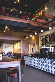 The interior of a modern coffee shop features a wooden and tile decor with a mix of industrial and cozy elements. The seating area includes several tables and chairs, and customers can be seen sitting and enjoying their drinks. Pendant lights hang from the ceiling, adding a warm ambiance. A wooden mezzanine level with additional seating is visible, overlooking the main floor. The counter area displays various coffee machines and accessories, with a patterned blue and white tile design on the front.