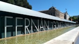 Large, bold letters spell out 'Fernán Gómez Centro Cultural' on a modern building facade. A shallow reflective pool stretches alongside the structure. In the background, classical architecture with a dome and a flag are visible. The setting is outdoors, with clear skies and greenery around.